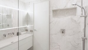 Um banheiro moderno com chuveiro em box de vidro, parede de mármore branco e toucador com espelho e pia.