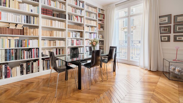 Uma biblioteca com estantes, uma mesa de vidro, cadeiras e vista para um edifício através de grandes janelas.