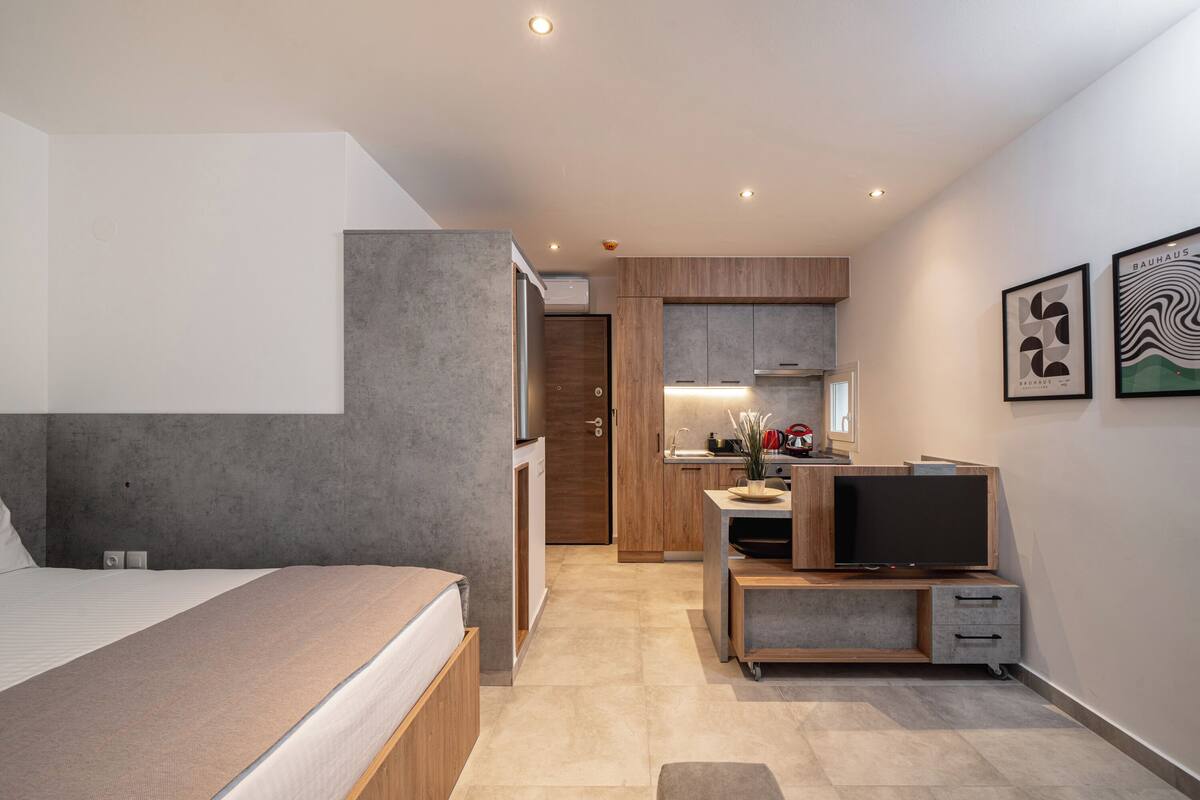 A modern hotel room with a bed, a desk with a flat-screen TV, a kitchenette, and a bathroom.