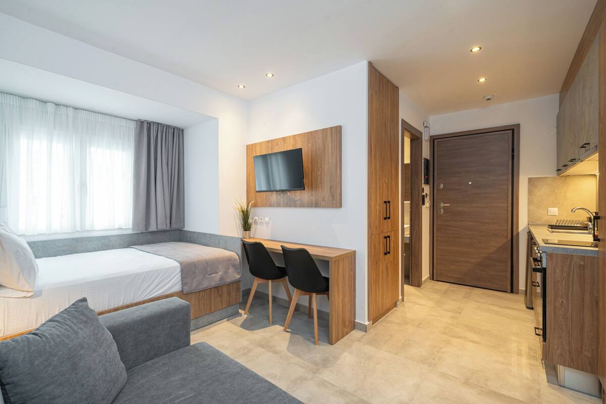 A modern hotel room with a bed, a desk, two chairs, a TV, a wooden wardrobe, and a kitchen area.