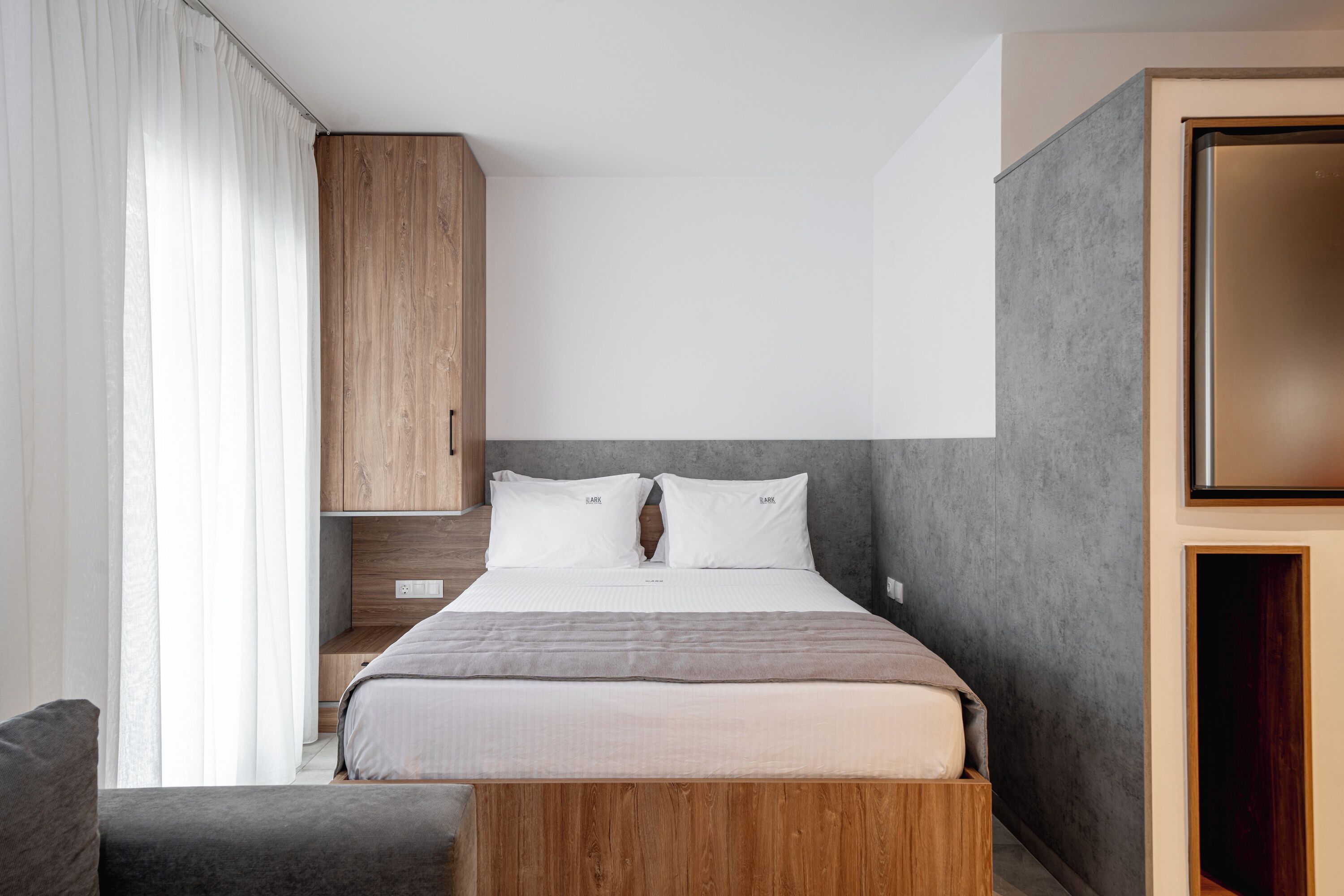 Un dormitorio moderno con cabecera de madera, cama con ropa de cama blanca, sofá gris y un espejo grande.
