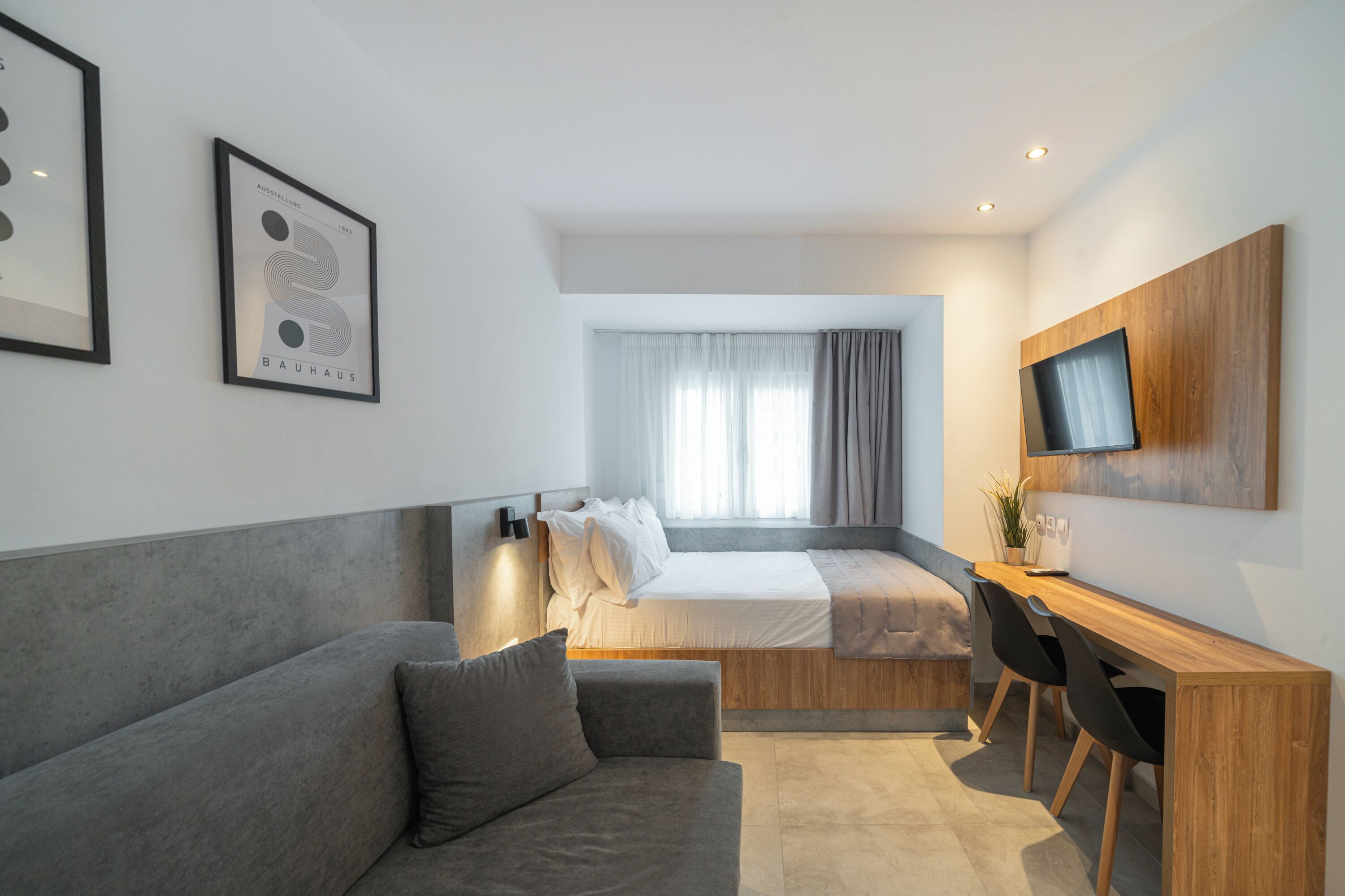 Una habitación de hotel moderna con un sofá gris, un escritorio de madera, una cama y un televisor instalado en la pared.