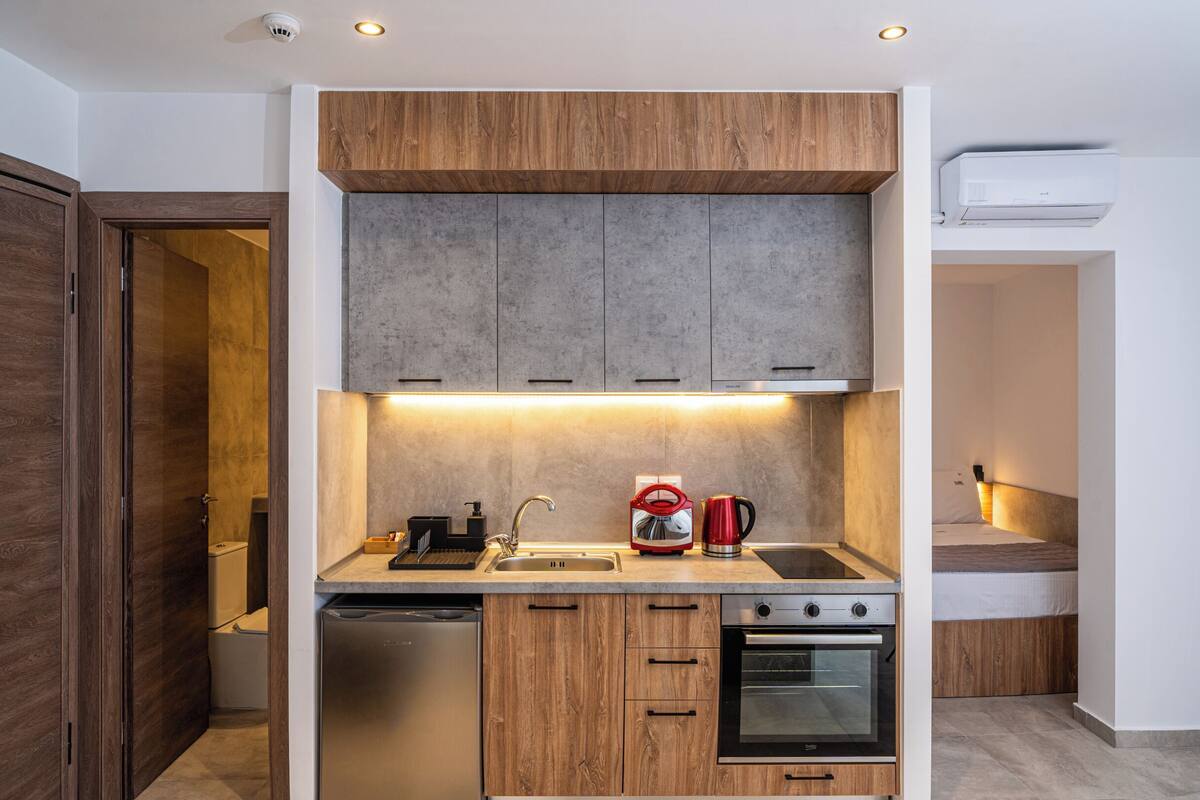 A compact kitchen with wooden cabinets, a stainless steel refrigerator, a red kettle, and an oven.