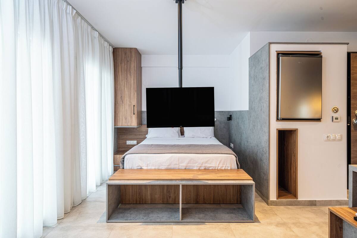 A modern hotel room with a bed, a flat-screen TV, a wooden bench, a wardrobe, and a door with a mirror.
