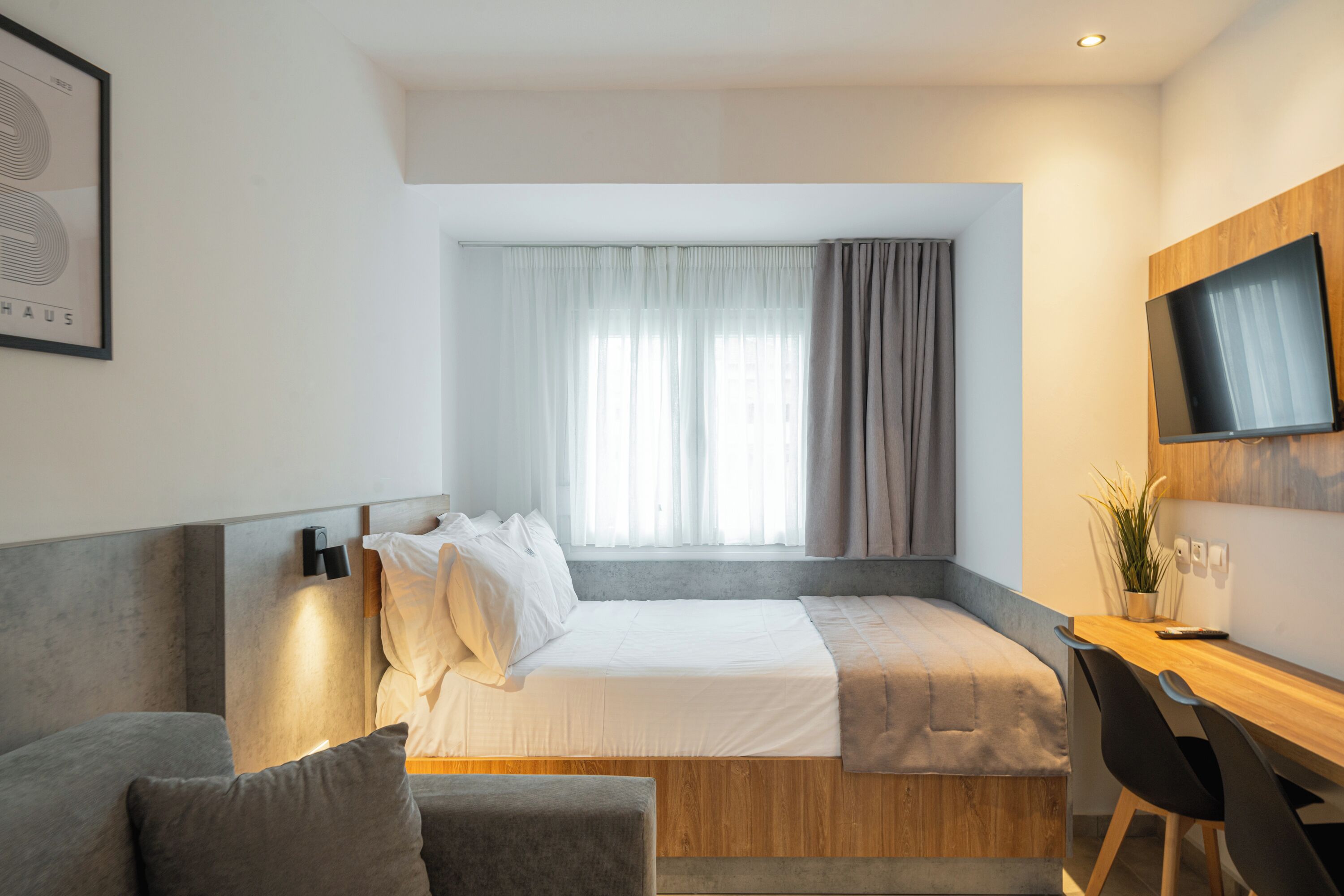 A modern hotel room with a bed, a desk, a TV, and a window with curtains.