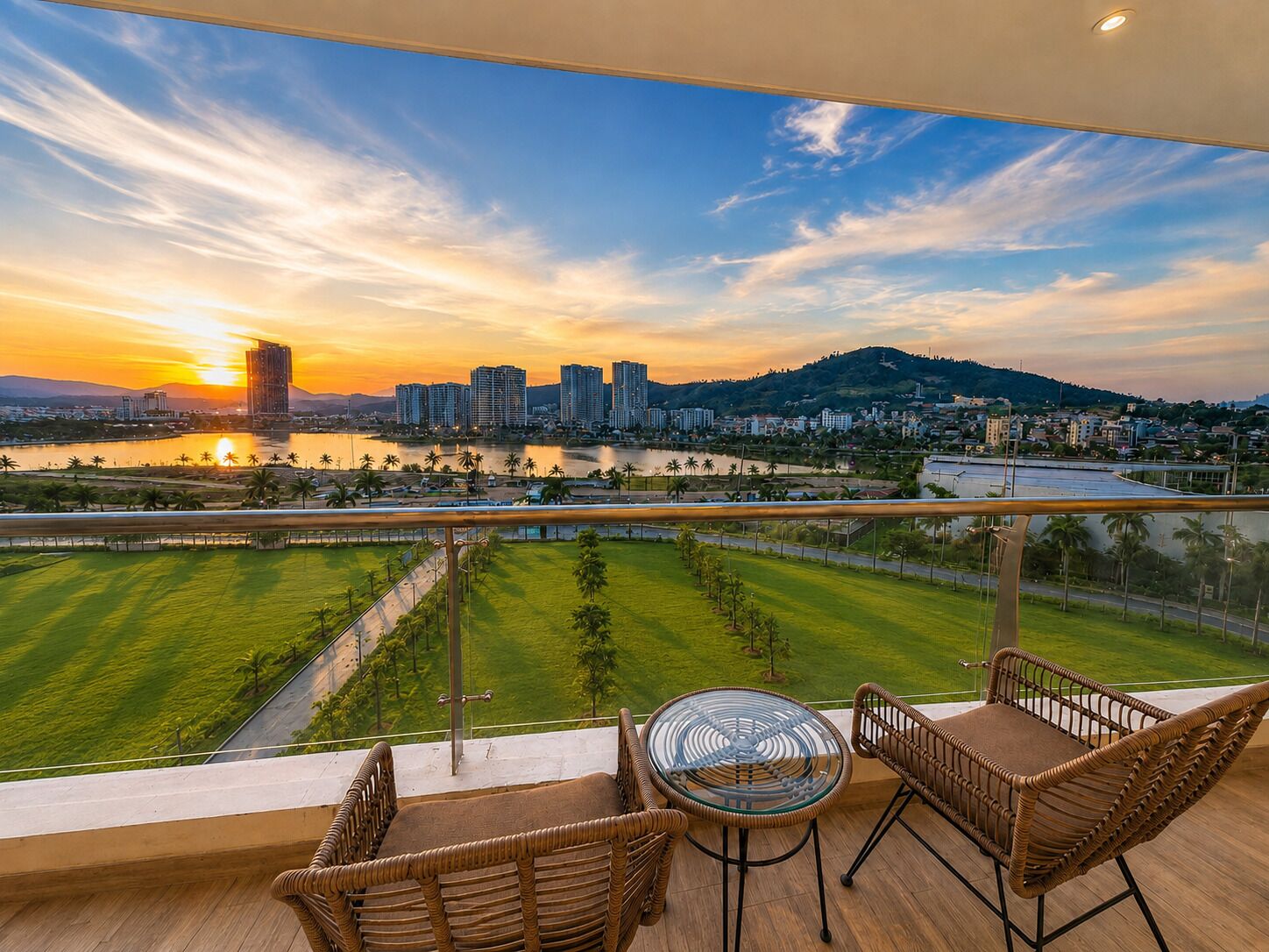 A balcony with wicker furniture, overlooking a cityscape with buildings and a river during sunset.