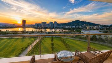 A balcony with wicker furniture, overlooking a cityscape with buildings and a river during sunset.