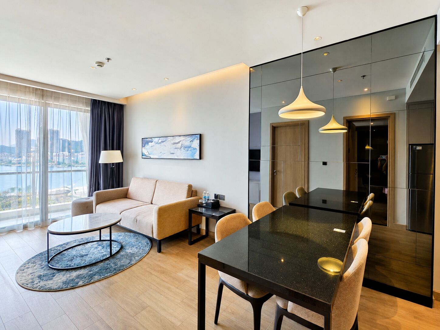 A modern hotel room with a sofa, dining table, and chairs, a large window with a view, and a painting on the wall.