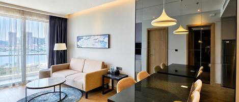 A modern hotel room with a sofa, dining table, and chairs, a large window with a view, and a painting on the wall.