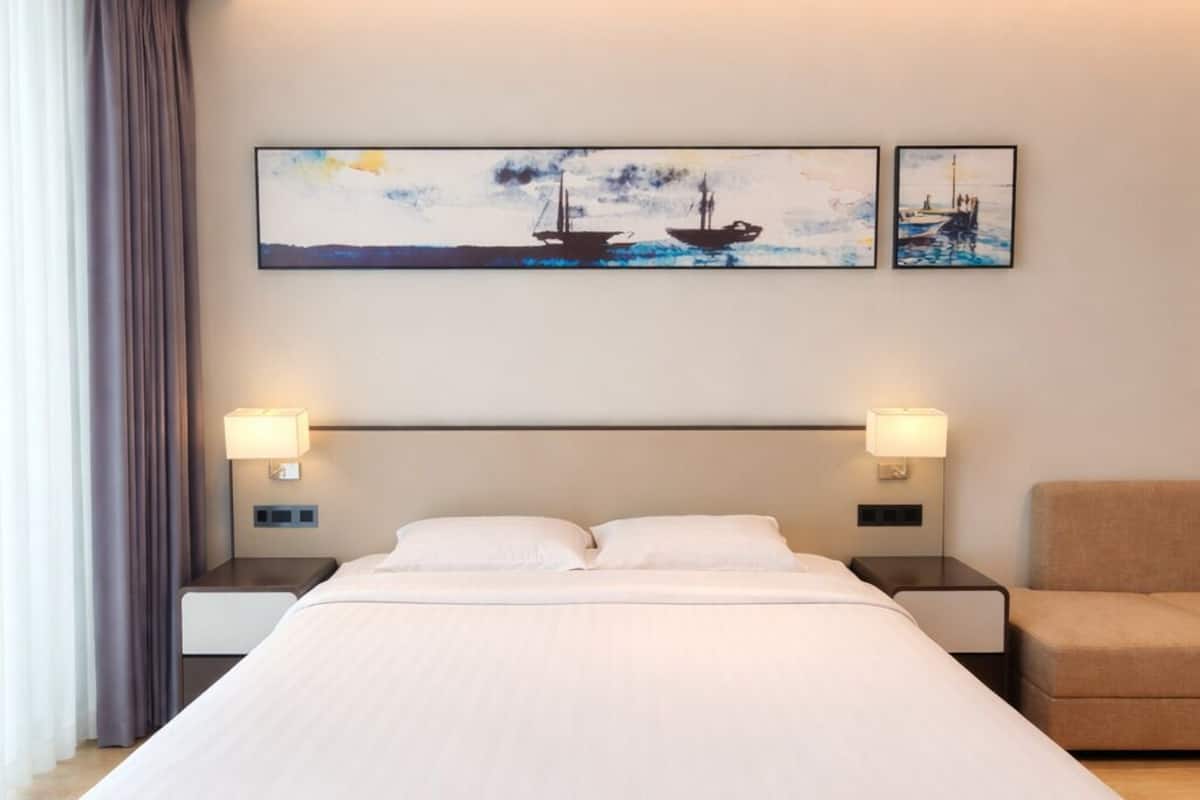 A large bed with a headboard, two bedside lamps, a chair, and two paintings on the wall.