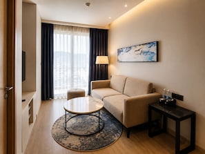 Une chambre dâhĂŽtel avec un canapĂ©, une table basse ronde, une lampe, un tableau et une table dâappoint avec des bouteilles dâeau.