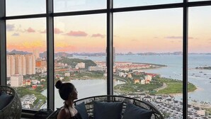 Une personne assise sur une chaise en osier, près d’une grande fenêtre donnant sur un paysage urbain au coucher du soleil.