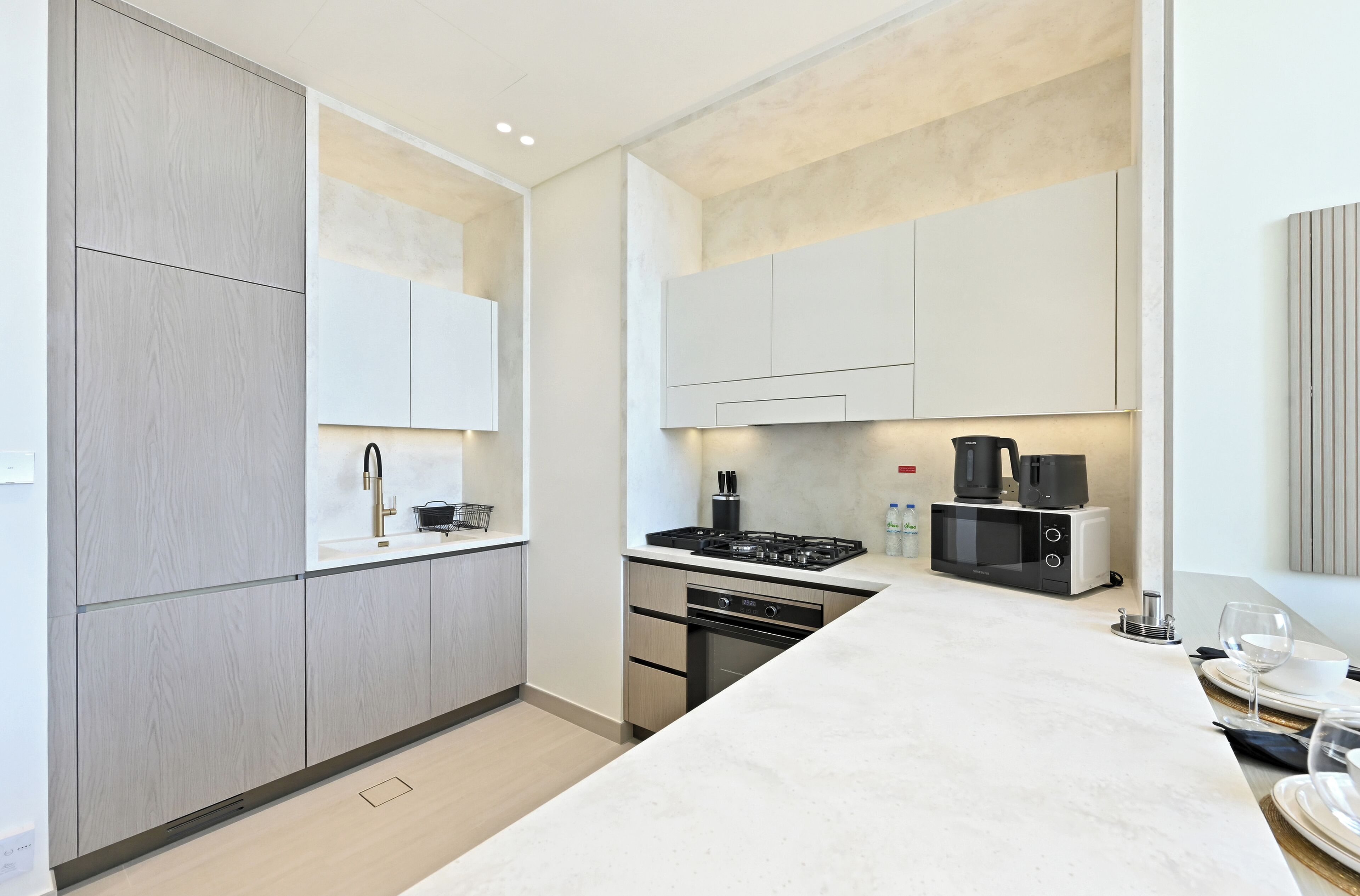 Une cuisine moderne avec des meubles en bois clair, un plan de travail blanc, une cuisinière, un four à micro-ondes et un coin repas avec une table dressée pour deux personnes.