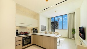 A modern kitchen with a central island, built-in oven, and microwave. There is a dining area with a white table and chairs, and a large window with a view of a construction site. - Bloomfields Iconic 1BR Binghatti Phanto (Dubai)