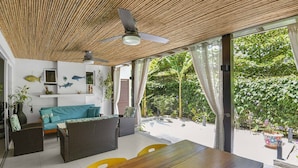 Ein Zimmer mit Deckenventilator, Korbmöbeln, einem Holztisch und Blick auf üppiges Grün außerhalb.