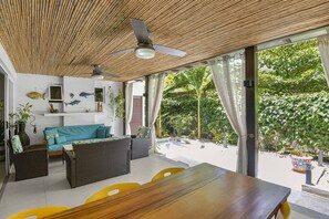 A room with a ceiling fan, wicker furniture, a wooden table, and a view of lush greenery outside. - Casa del Mar Tropical Sanctuary (Jacó)