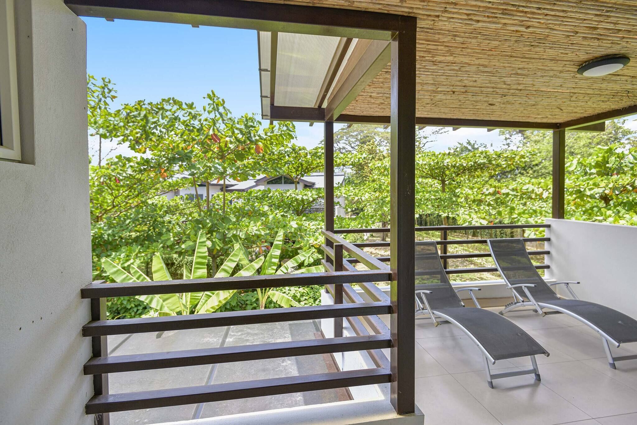 A covered outdoor seating area with metal railings and lounge chairs, overlooking a garden with trees and a building in the background.