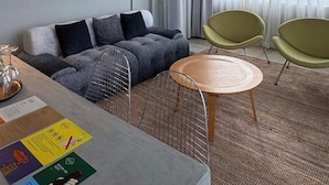 Een moderne woonkamer met een grijze bank, groene stoelen, een ronde houten tafel en een groot raam met dunne gordijnen.