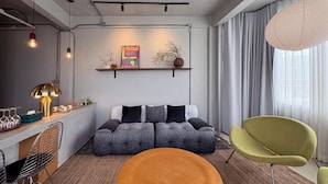 Een moderne woonkamer met een bank, een ronde houten tafel en een groene stoel. Links staat een plank met decoratieve objecten en een lamp.