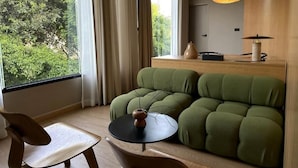 Een moderne kamer met een groene bank, een kleine tafel met een vaas en uitzicht op bomen door grote ramen.