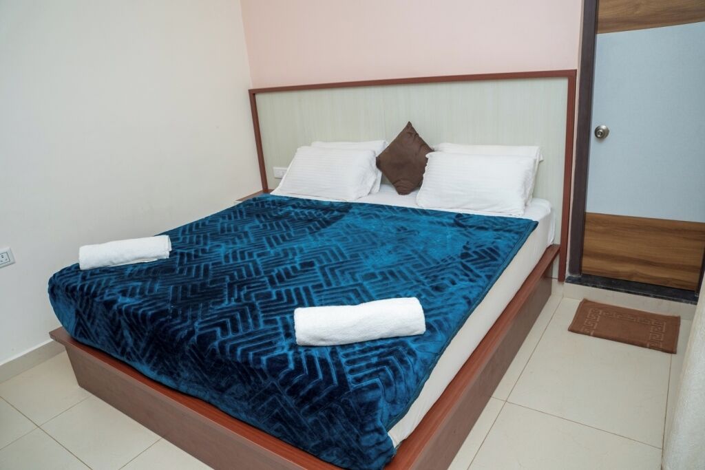 A neatly made bed with a patterned blue bedspread and white pillows, a wooden headboard, and a brown doormat on the floor.