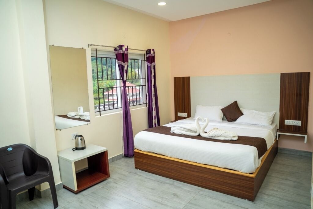A hotel room with a wooden bed, a small bedside table with a kettle, a chair, and a window with purple curtains.