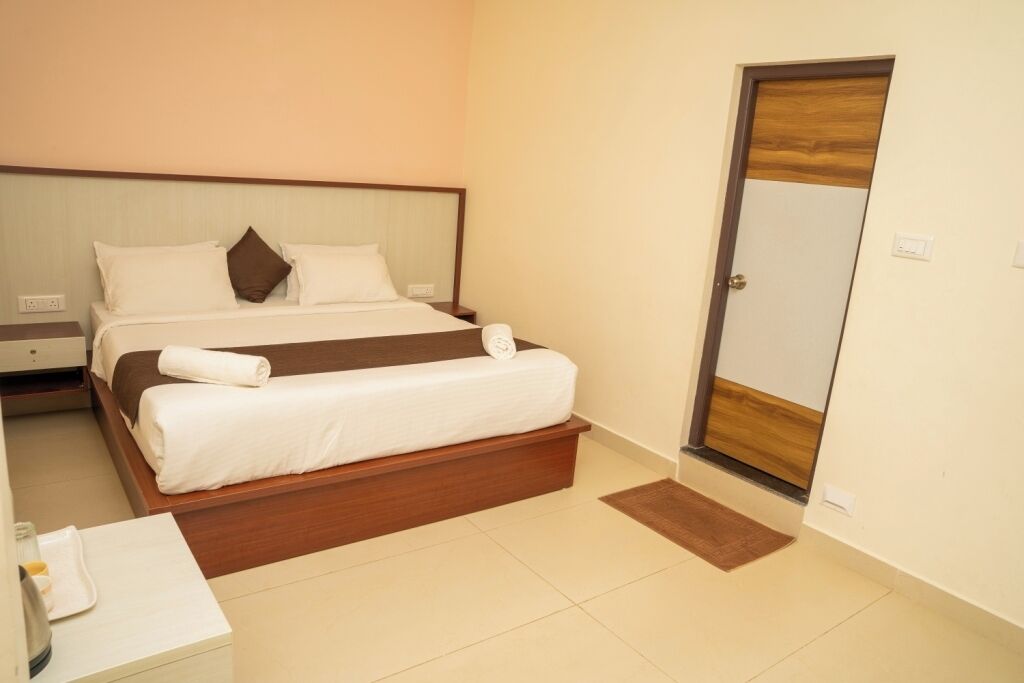 A neatly made bed with white linens and a brown throw, a wooden nightstand with a lamp, and a door with a wooden panel.
