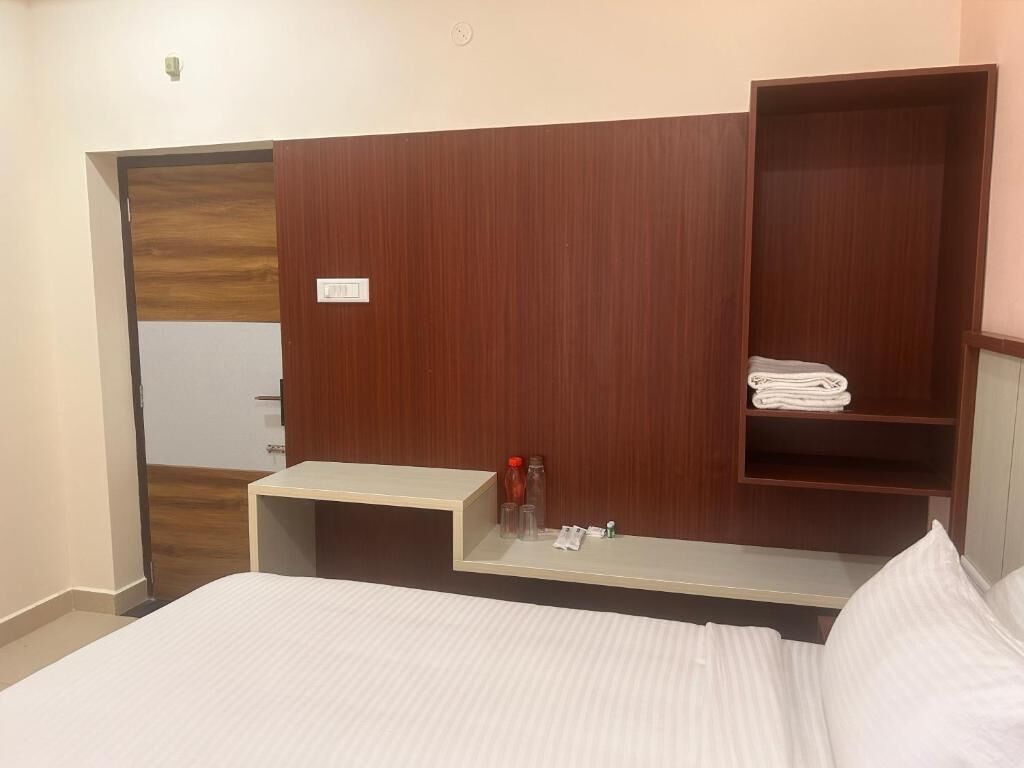 A hotel room with a bed, a wooden headboard, a small table with toiletries, and a shelf with folded towels.