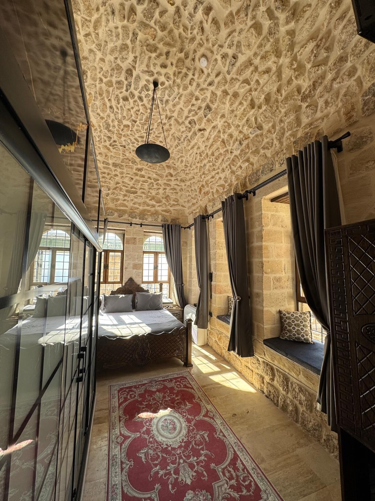 Une chambre avec un plafond en pierre, un grand lit recouvert de plusieurs coussins, un tapis rouge à motifs et un coin fenêtre agrémenté d’un coussin.