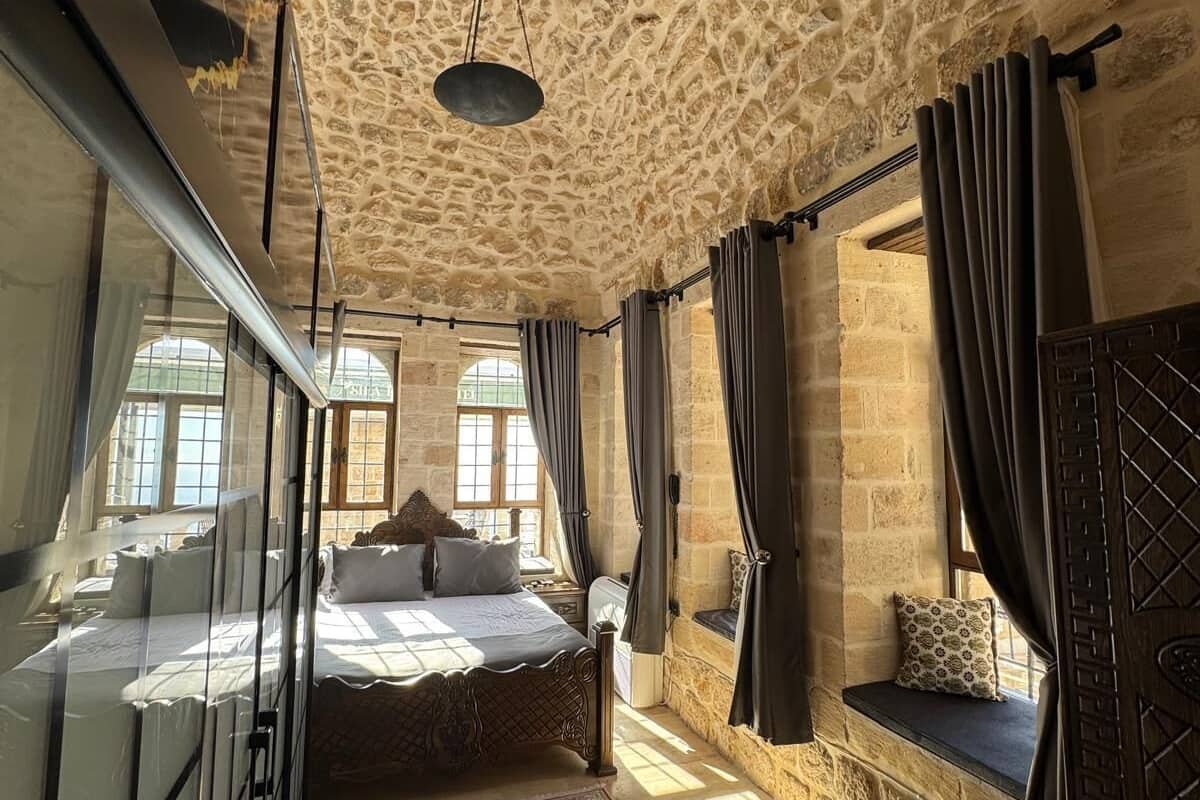 Un dormitorio con techo de piedra, una cama grande con muchas almohadas, una alfombra roja con estampado y un asiento junto a la ventana con cojín.