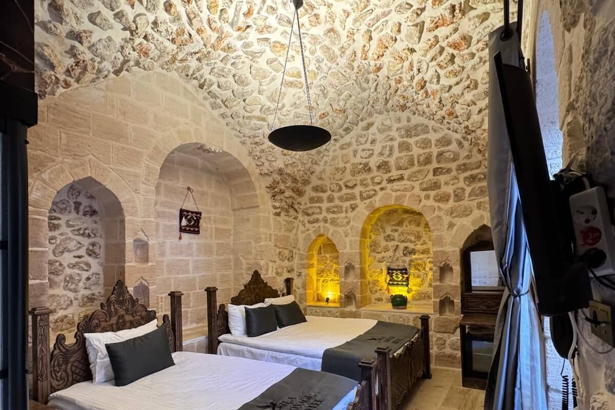 Una habitación con paredes de piedra, dos camas con cabeceros de madera, un tapete rojo con estampado y un aire acondicionado de pared.