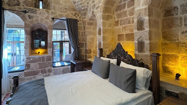 Un dormitorio con paredes de piedra, una cama grande, un candelabro colgante y una ventana con cortinas.