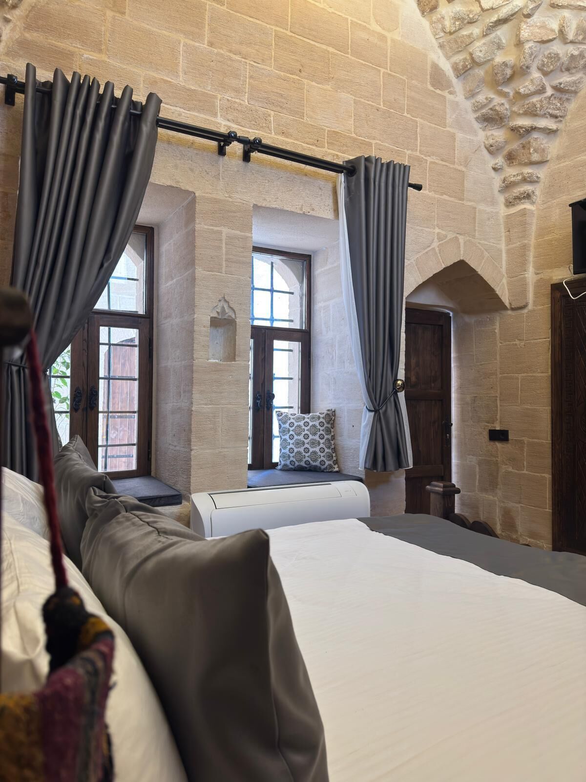 Een slaapkamer met een stenen muur, een bed, een bankje en een raam met gordijnen.