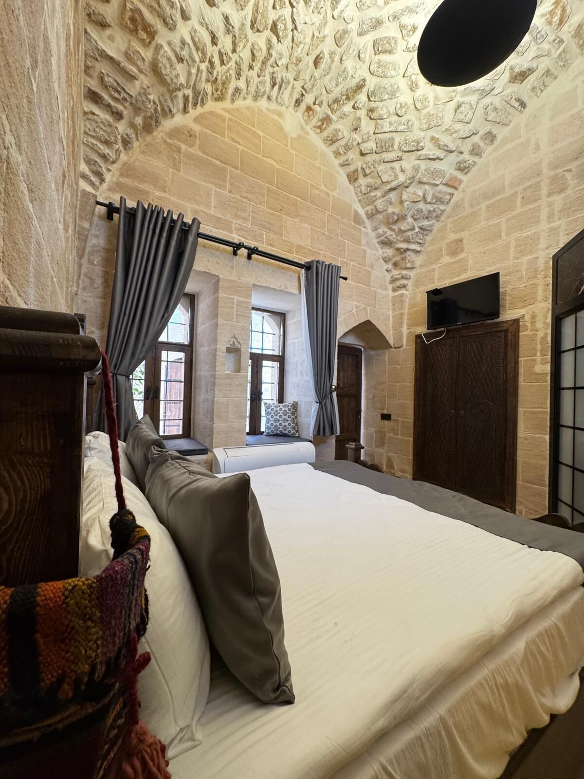 Une chambre avec un mur en pierre, un lit avec du linge de lit blanc, des rideaux gris et une tête de lit en bois.