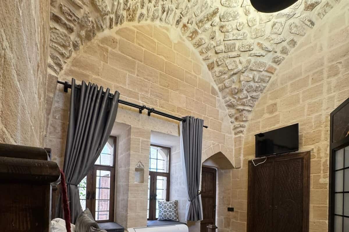 Un dormitorio con pared de piedra, cama con ropa de cama blanca, cortinas grises y cabecero de madera.