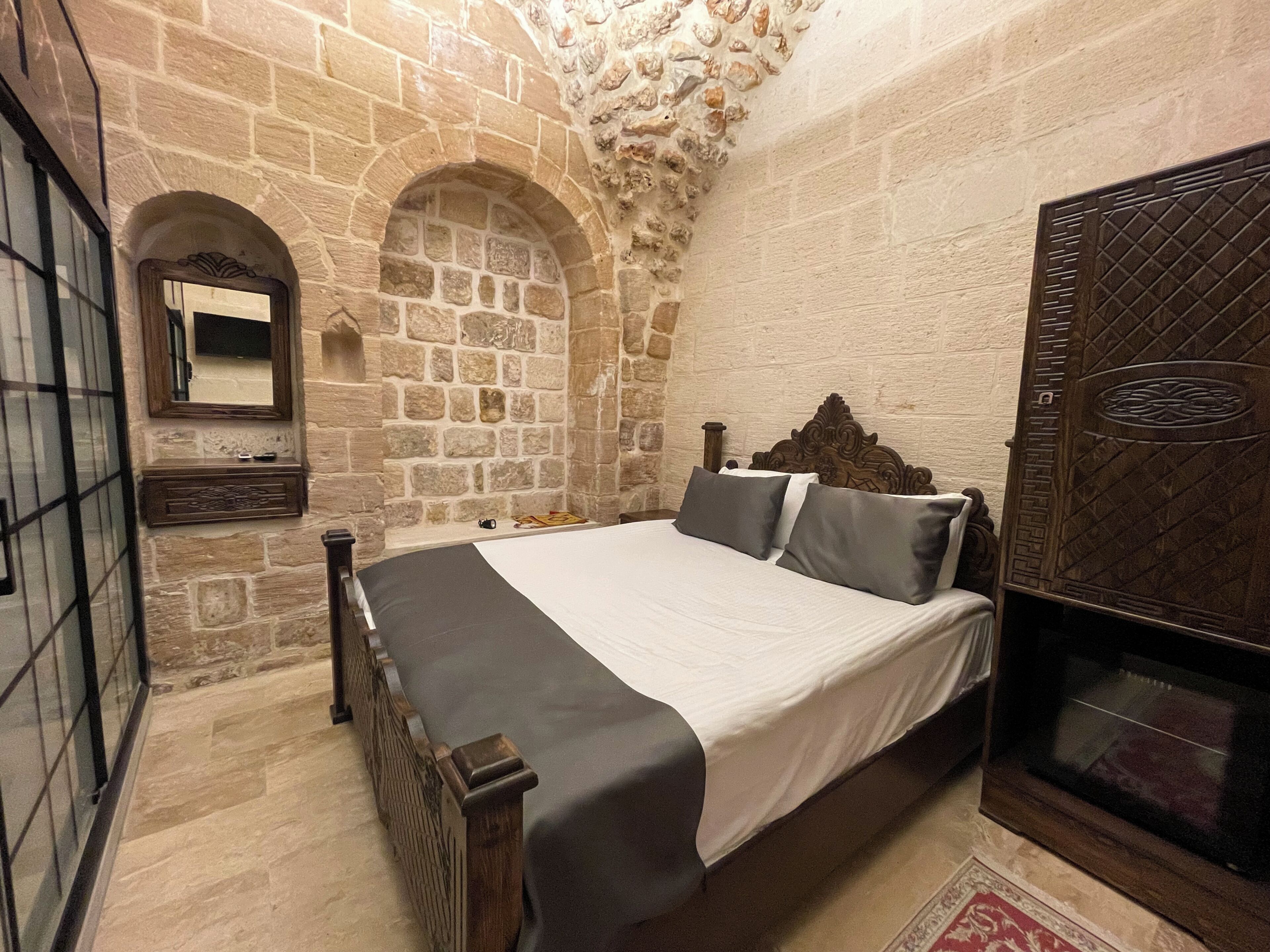 Une chambre avec un mur en pierre, un lit en bois avec une tête de lit, une table de chevet, un miroir et une armoire.