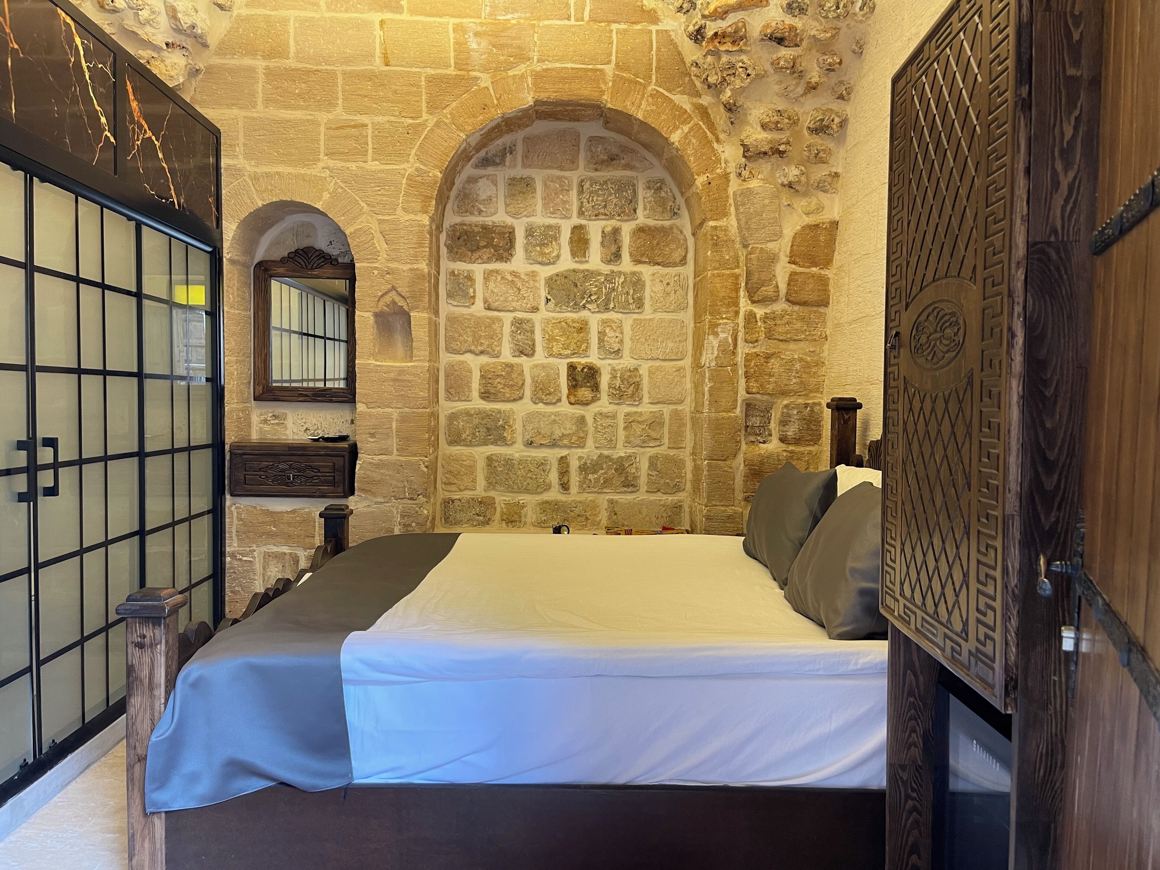 Une pièce avec un mur en pierre, un lit avec un matelas blanc et des oreillers gris, une porte en bois ornée de sculptures complexes, et une petite table de chevet en bois avec un miroir et un tableau encadré.