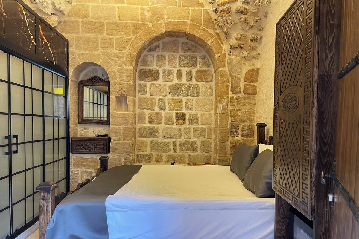 Una habitación con pared de piedra, una cama con colchón blanco y almohadas grises, una puerta de madera con tallas intrincadas y una pequeña mesita de noche con un espejo y un cuadro enmarcado.