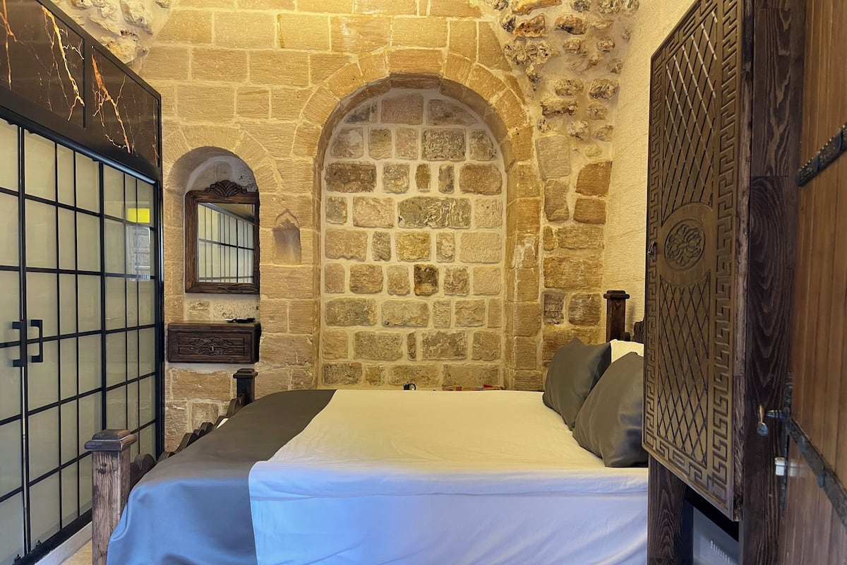 Une pièce avec un mur en pierre, un lit avec un matelas blanc et des oreillers gris, une porte en bois ornée de sculptures complexes, et une petite table de chevet en bois avec un miroir et un tableau encadré.