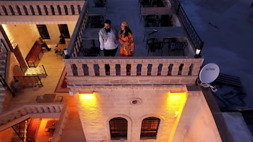 A couple standing on a balcony overlooking a courtyard with a stone building, outdoor seating, and a satellite dish.