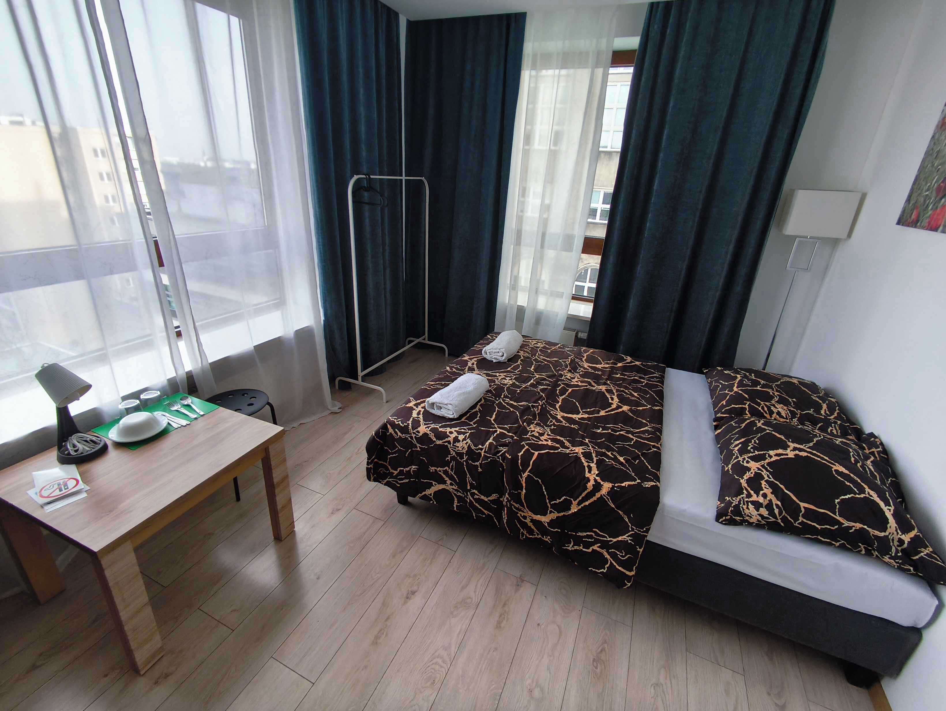 A hotel room with a bed, a desk, a chair, a window with curtains, and a view of buildings outside.