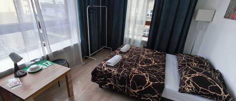 A hotel room with a bed, a desk, a chair, a window with curtains, and a view of buildings outside.
