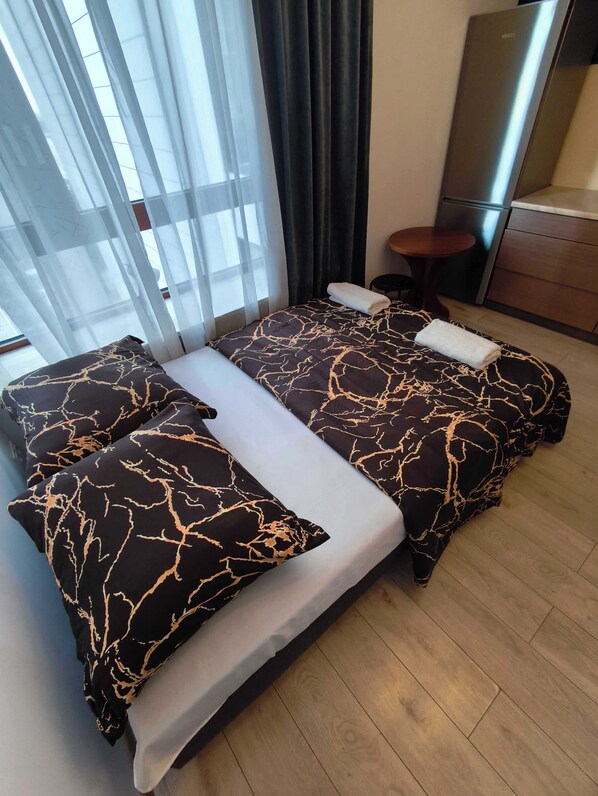 Un lit double recouvert d’une couvre-lit à motifs noirs et dorés, un oreiller blanc et une serviette pliée posés sur le lit. On aperçoit une petite table en bois à côté du lit, une chaise de couleur sombre et, au fond, une petite cuisine moderne.
