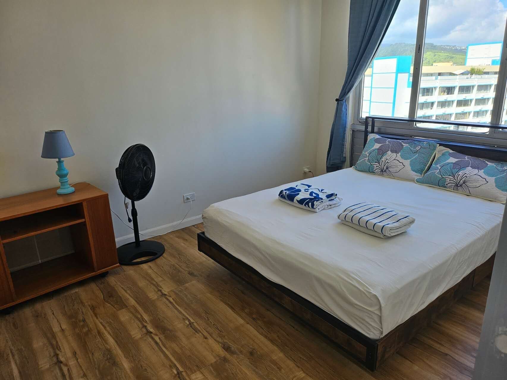 Ein Schlafzimmer mit einem Bett, einem Nachttisch mit Lampe, einem Standventilator und einem Fenster mit Vorhängen.