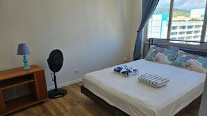 Ett sovrum med en säng, ett nattduksbord med en lampa, en golvfläkt och ett fönster med gardiner.