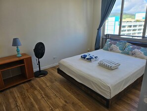 Ein Schlafzimmer mit einem Bett, einem Nachttisch mit Lampe, einem Standventilator und einem Fenster mit Vorhängen.
