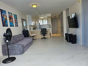 Een woonkamer met een bank, een ventilator, een televisie en een keuken op de achtergrond.
