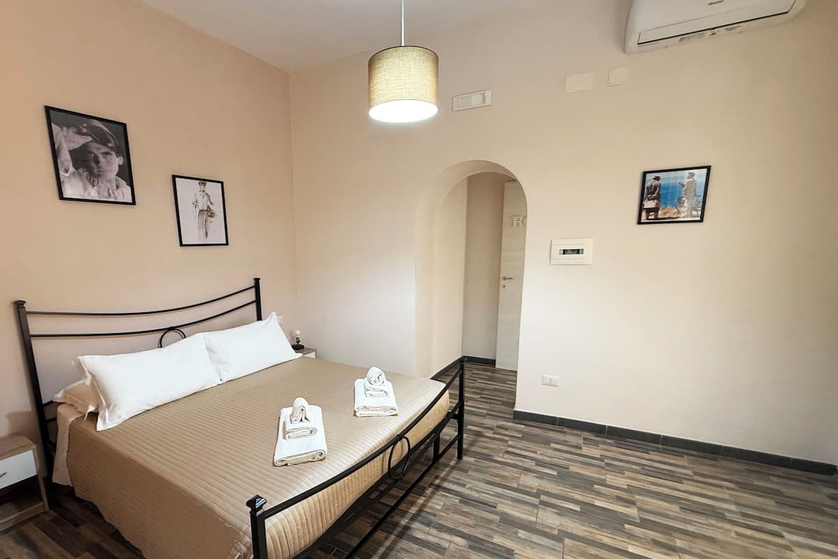 A neatly arranged bedroom with a bed, a hanging light fixture, and a door leading to another room.