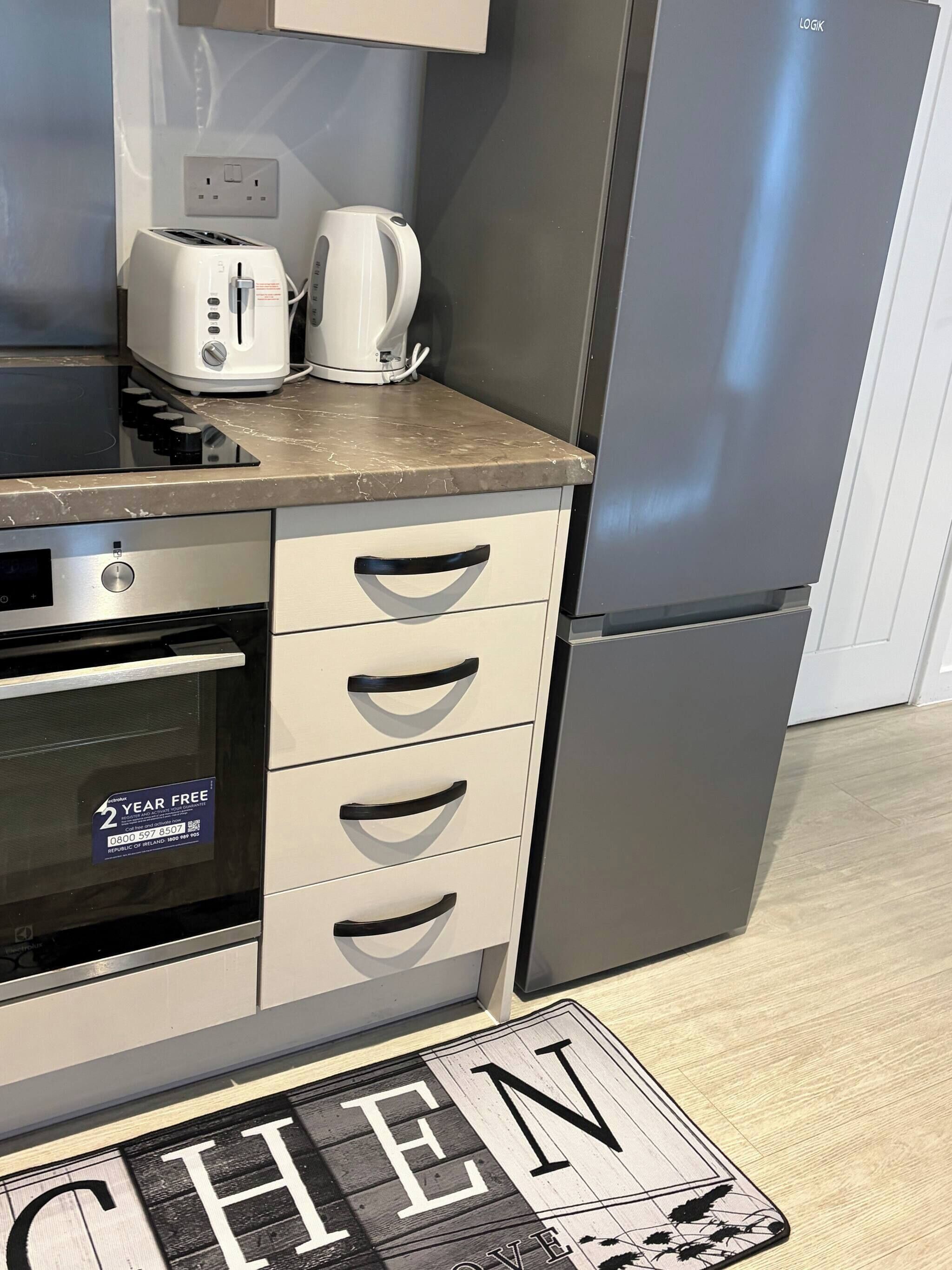 Eine Küche mit ein eingebautem Ofen, einem Toaster und einem Wasserkocher auf der Arbeitsplatte. Ein Edelstahl-Kühlschrank und ein Holzboden sind ebenfalls vorhanden.