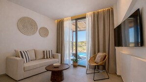A living room with a sofa, a round coffee table, a wicker chair, a TV, and a large window with curtains. - Ramnus Karpathos Suites (Karpathos)
